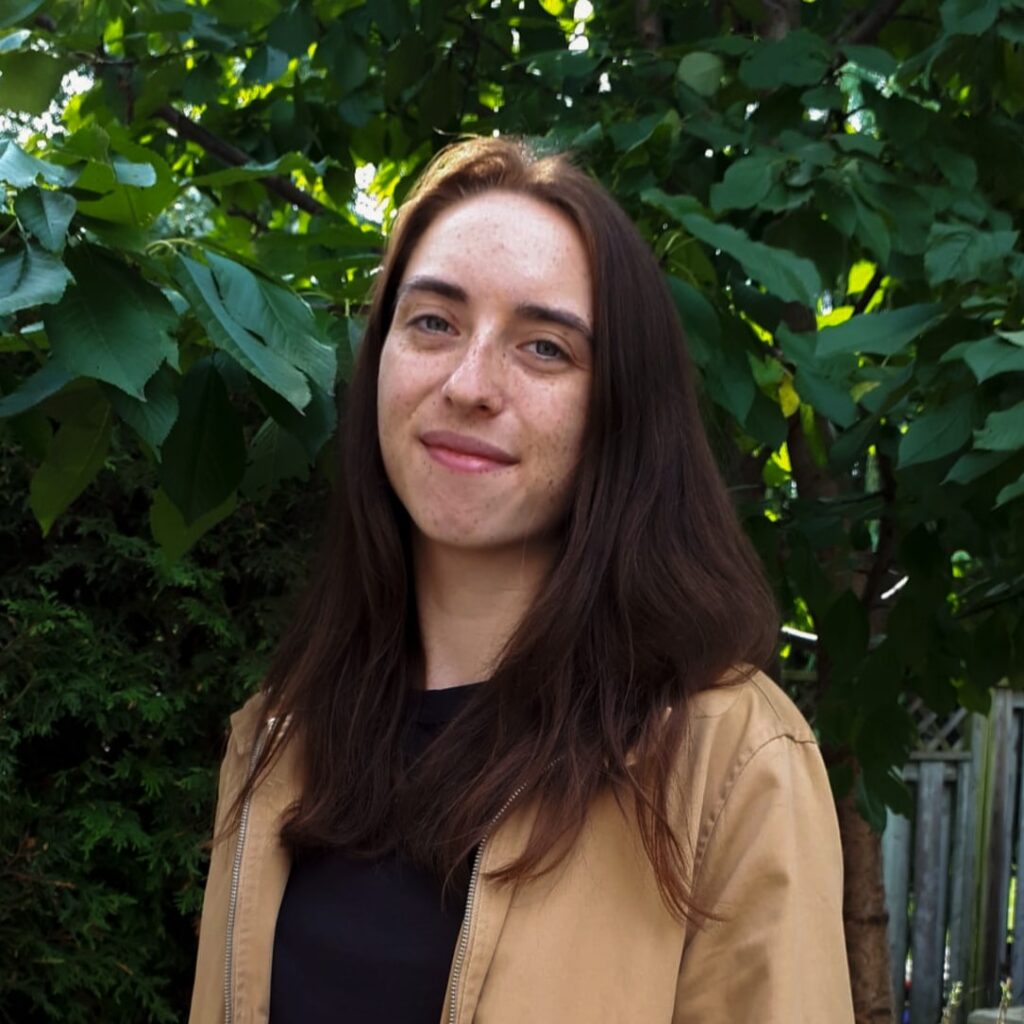 Young woman with brown hair photographed outdoors in front of foliage.