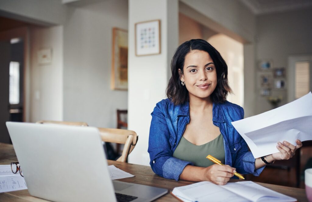 A young woman sitting at her home office in front of a laptop and writing in a notebook.