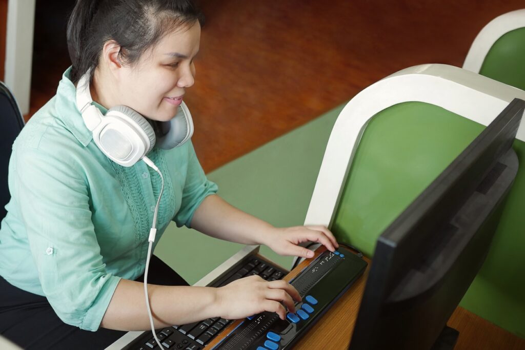 A blind young woman sits at a computer desk and uses an adaptive brailled keyboard in front of a computer monitor.